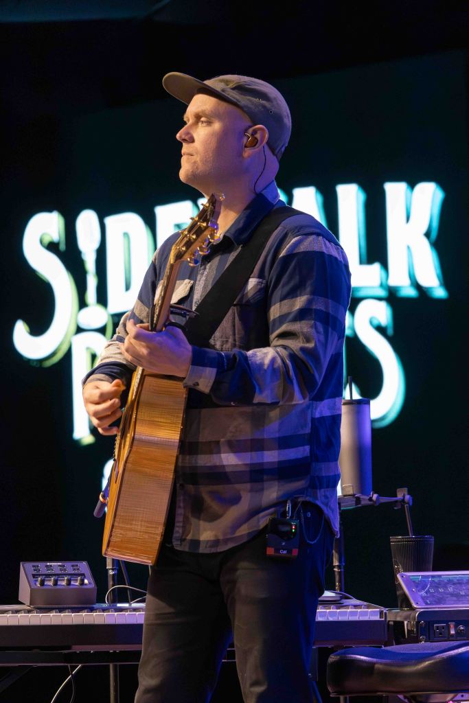 A musician playing an acoustic guitar on stage, wearing a plaid shirt and a cap, with audio equipment and a keyboard in the background.