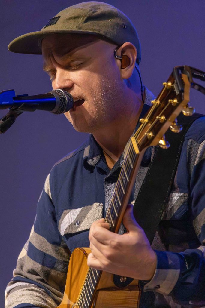 Musician performing live while singing into a microphone and playing an acoustic guitar, wearing a cap and a striped shirt.