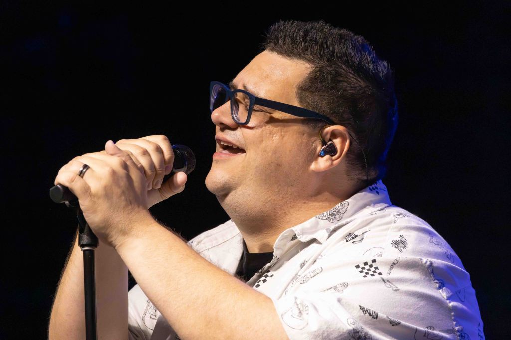 A male singer passionately performing on stage while holding a microphone, wearing a patterned shirt and glasses.