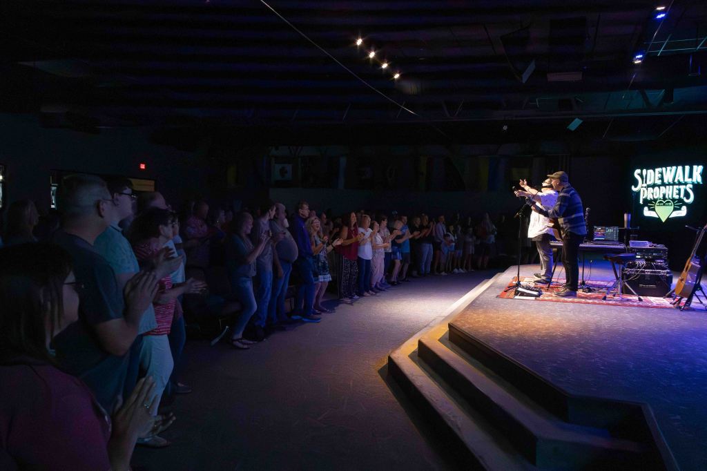 A live performance by the musical group Sidewalk Prophets in a dimly lit venue, with an audience clapping and enjoying the show.