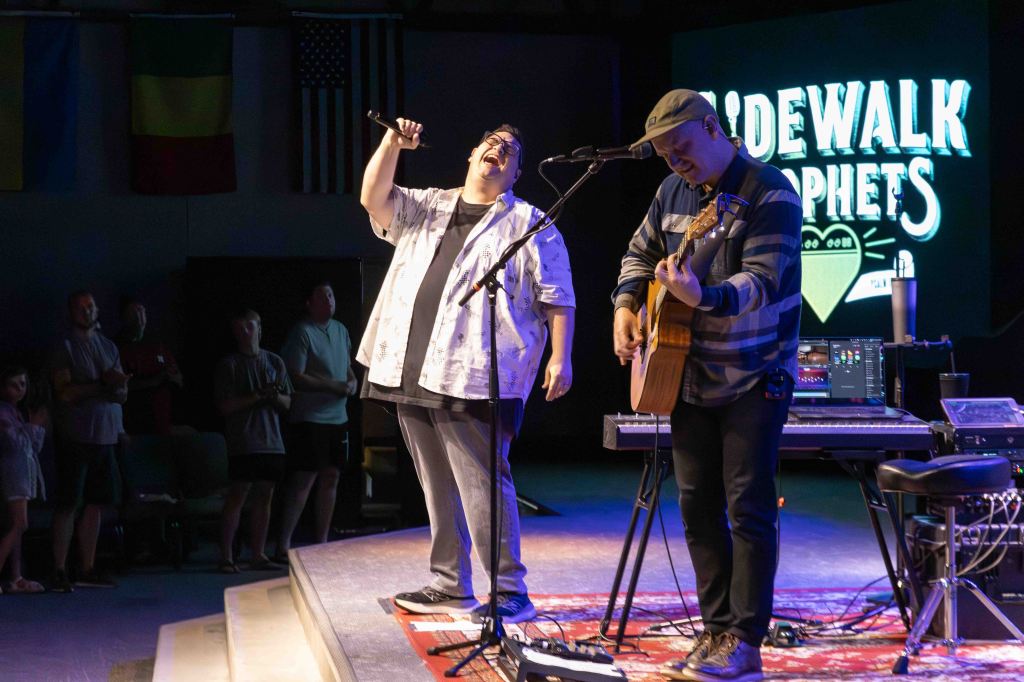 Two male performers on stage during a live music event, one singing into a microphone and the other playing an acoustic guitar, with an audience visible in the background.