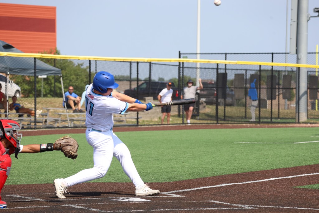 A baseball player in a white uniform with blue accents swings a bat at a pitch on a baseball field, while a catcher in red gear prepares to catch the ball.