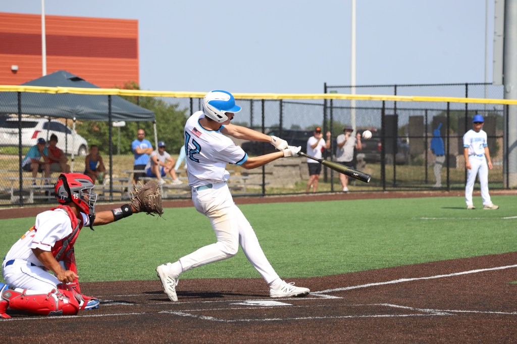 A baseball player with the number 12 swings a bat at a pitch while a catcher in red gear prepares to receive the ball during a game on a sunny day.