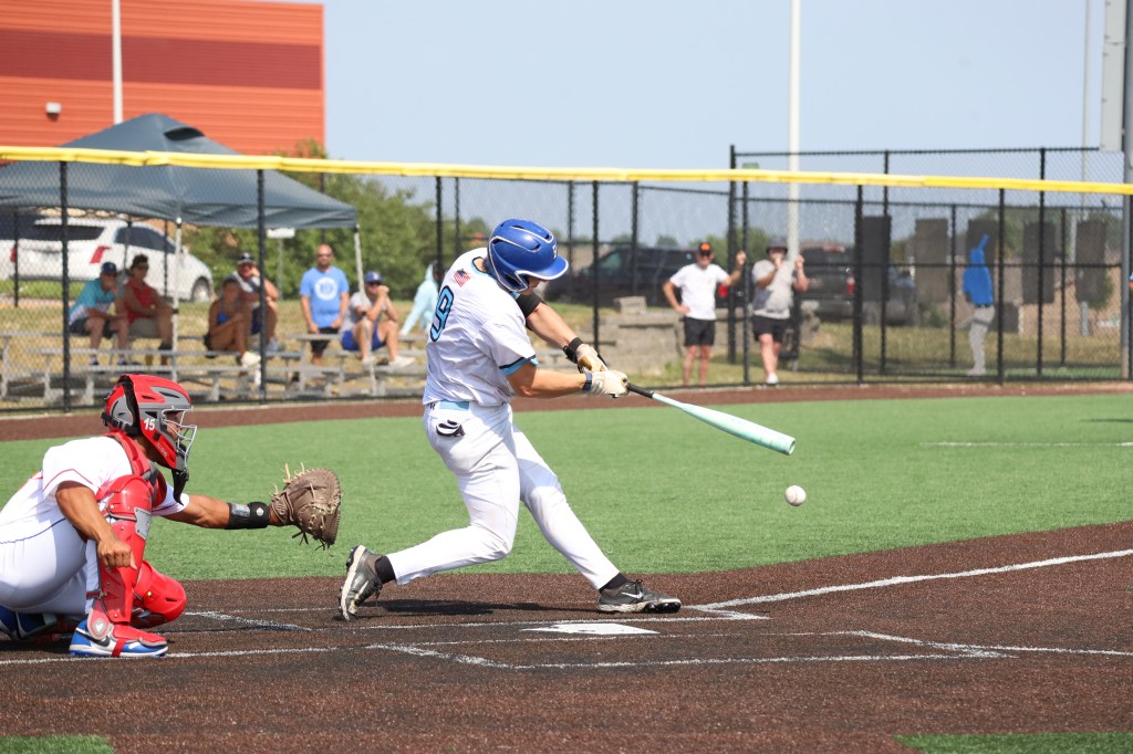 A baseball player in a white uniform swings at a pitched ball, while a catcher in red crouches behind home plate. Spectators are visible in the background.