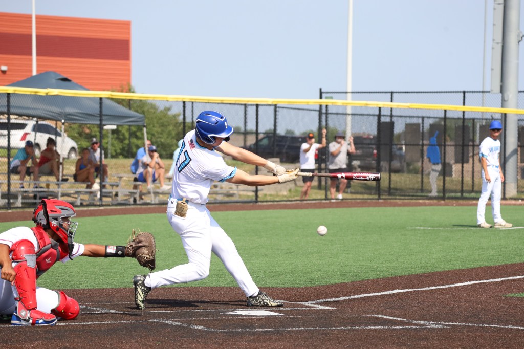 A young baseball player in a blue jersey swings a bat at a pitch during a game, with a catcher in red gear poised behind him and spectators in the background.