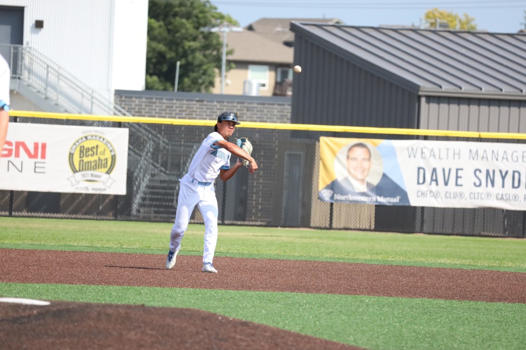 A young baseball player fields a ball and prepares to throw it during a game, with a field and spectators in the background.