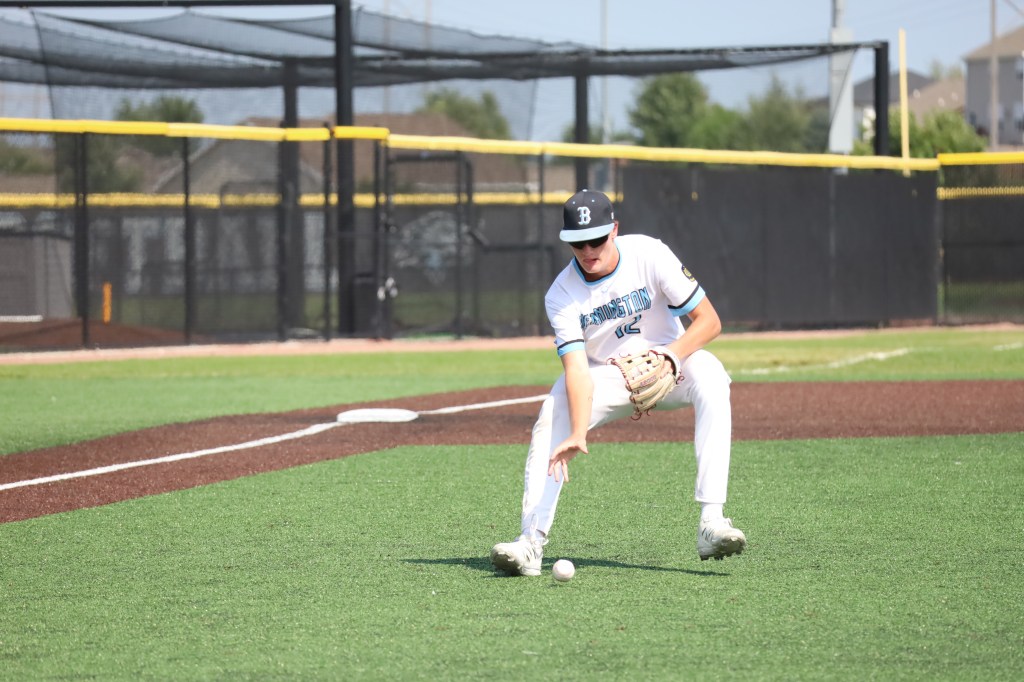 A young baseball player in a white uniform crouches to field a ground ball on a baseball diamond.