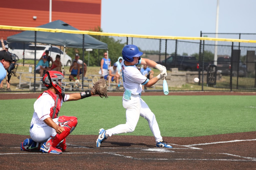 A baseball player in a blue helmet and white uniform swings at a pitch while the catcher in red gear prepares to receive the ball. The scene takes place on a sunny day at a baseball field.