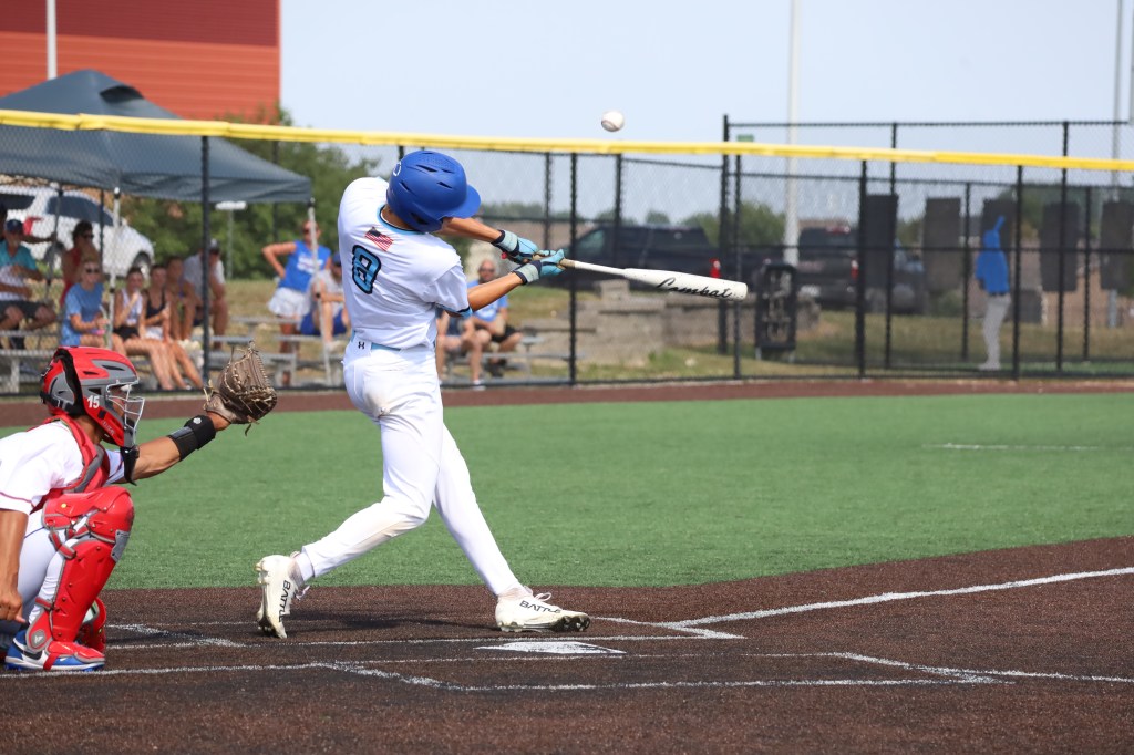 A young baseball player in a light blue uniform swings a bat at a pitch during a game, while a catcher in red gear prepares to catch the ball.