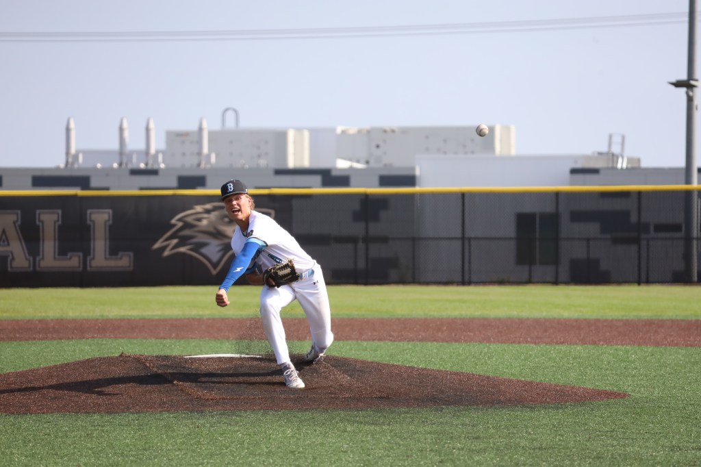A baseball pitcher in mid-throw on a field, wearing a white and blue uniform with a cap, showing an expressive facial expression.