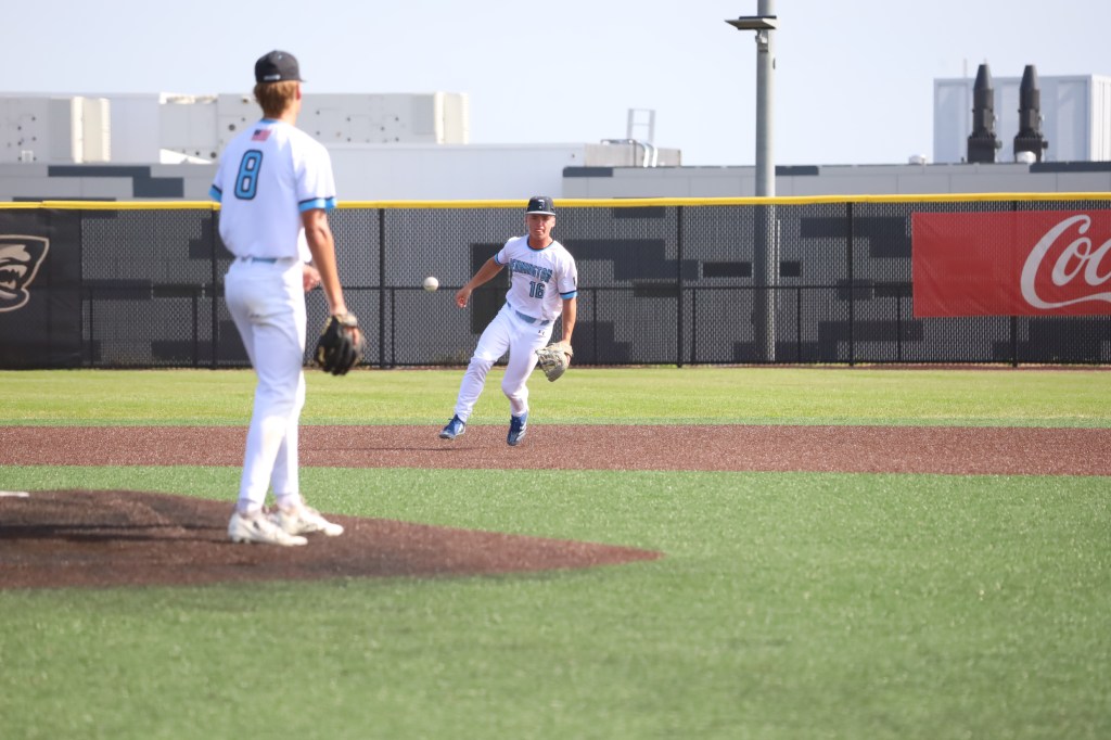 A baseball player in a white uniform is catching a ball while another player in the background prepares to pitch on a baseball field.
