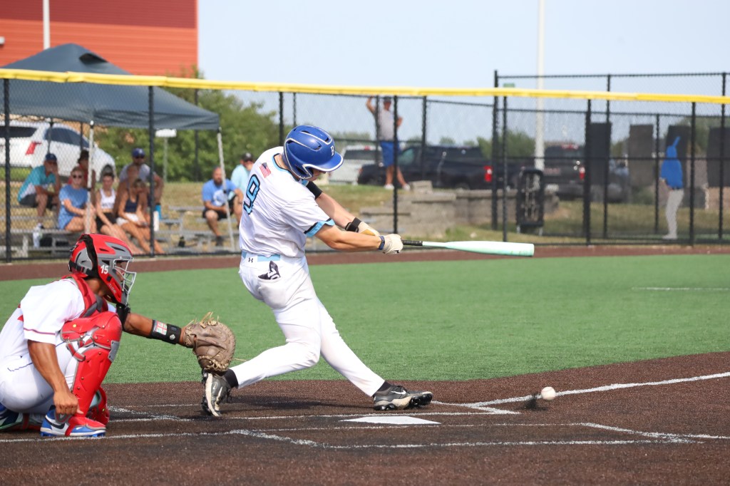 A baseball player swings at a pitch while a catcher crouches behind home plate, with spectators visible in the background.