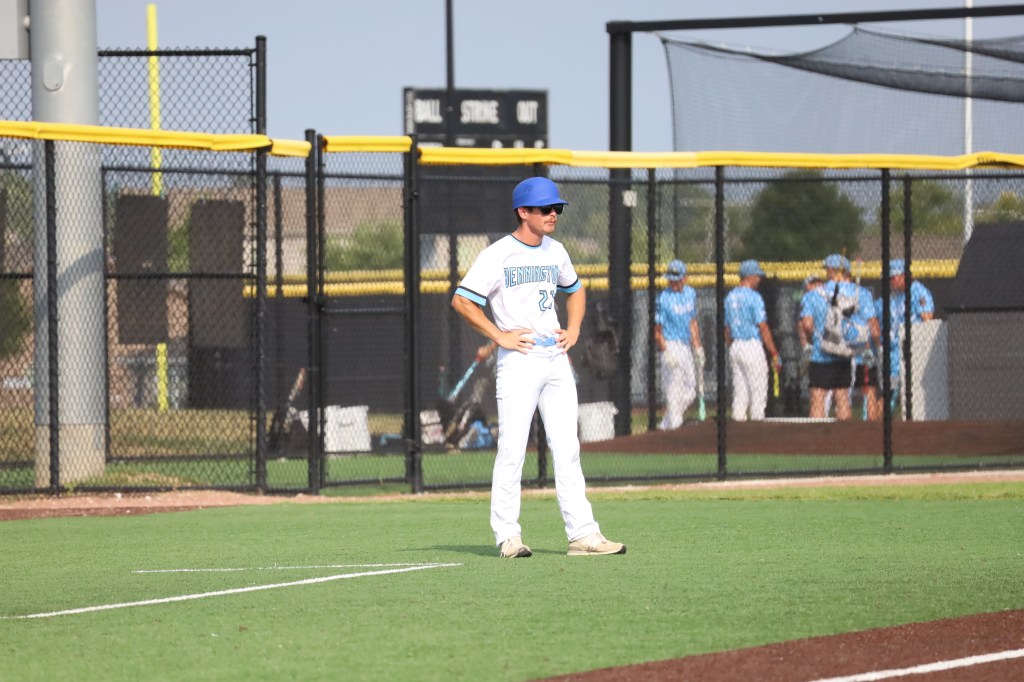 A baseball player stands on the field, wearing a blue helmet and white uniform, with hands on hips, observing teammates in the background.