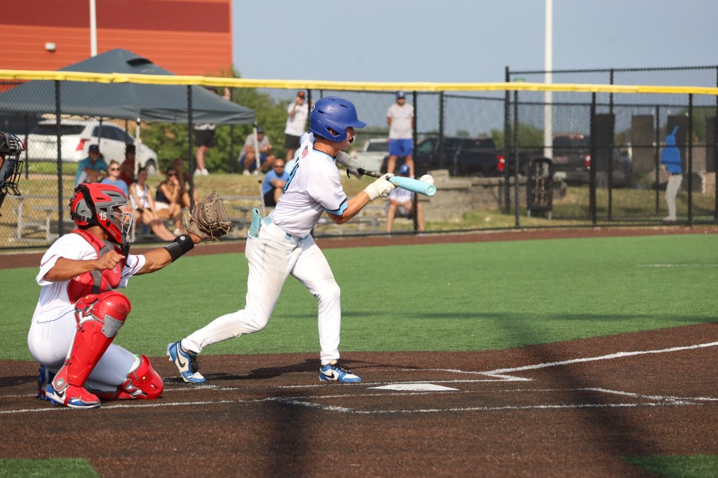 A young baseball player swings a bat while the catcher prepares to receive the pitch, with spectators in the background.