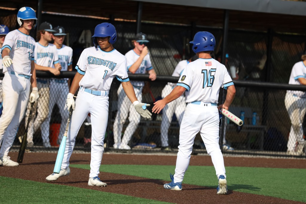 Two young baseball players in uniforms greeting each other on the field, with teammates in the background.