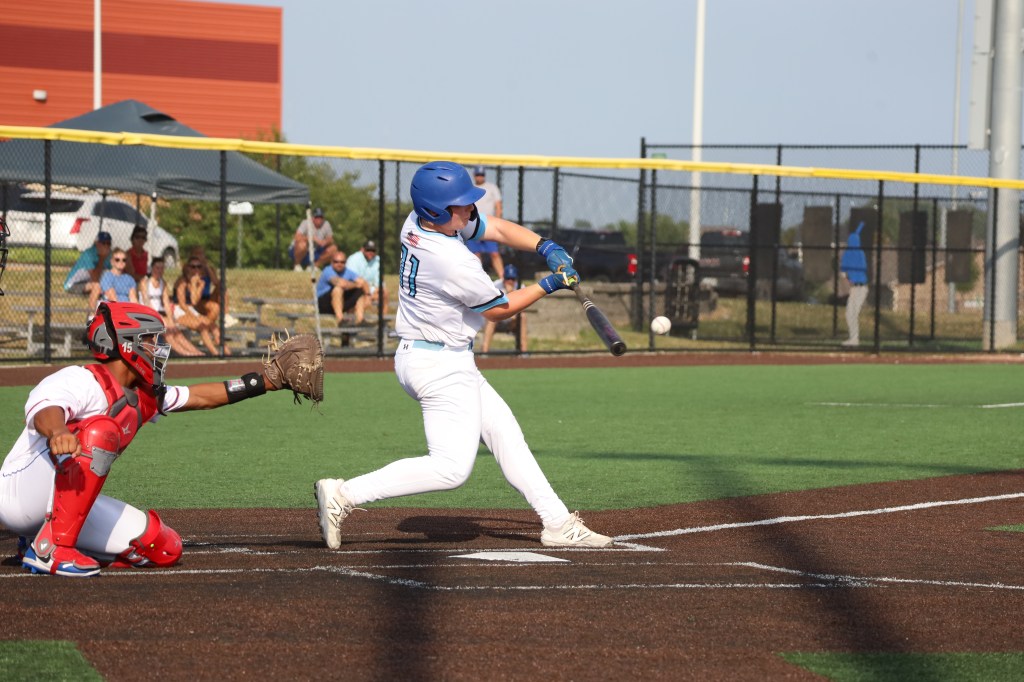 A baseball player in a blue cap swings a bat at a flying baseball while a catcher in red gear prepares to catch. The scene takes place on a green turf field with spectators in the background.