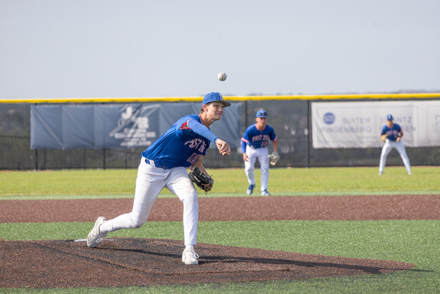 A baseball pitcher in a blue jersey throwing a pitch on the mound, with two fielders in the background on a baseball field.