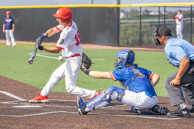 A baseball player in a red helmet and jersey swings at a pitch while a blue catcher crouches behind home plate. An umpire in a blue shirt observes the play.