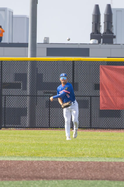 A baseball player in a blue jersey throws a ball while on the field, with a black fence and a colorful background visible.