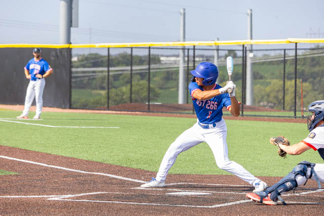 A youth baseball player in a blue jersey and cap prepares to swing at the pitch, with a catcher in gear ready to receive the ball and a teammate on deck in the background.