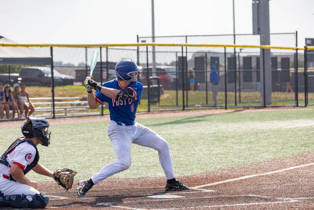 A young baseball player in a blue jersey swings a bat while batting, with a catcher in a black and white uniform crouched behind home plate.