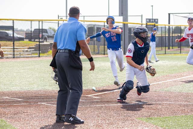 A youth baseball game scene showing a player in a blue jersey throwing a ball while another player in catcher's gear attempts to evade a tag. An umpire watches the play closely.