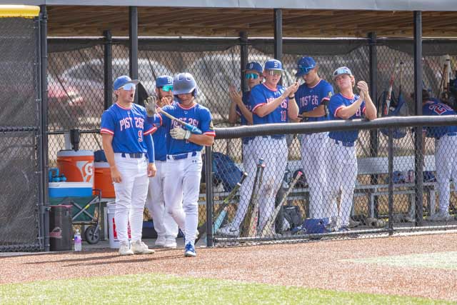 Baseball team members in uniforms standing in the dugout, with some players preparing to play and others watching the game.