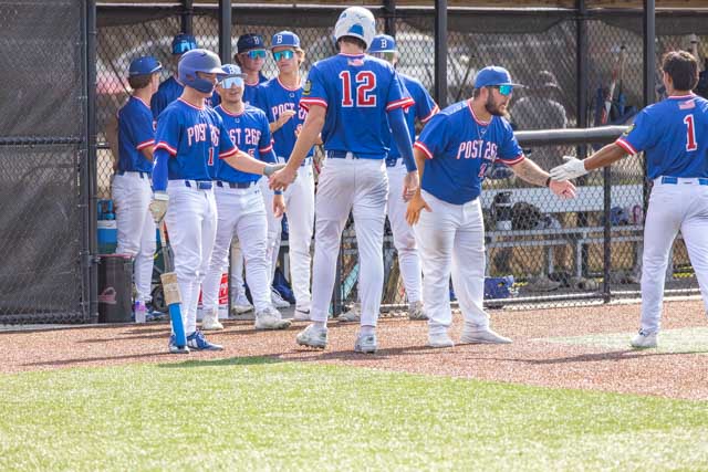 A group of baseball players in blue jerseys with 'Post 26' visible, celebrating and high-fiving at the dugout during a game.
