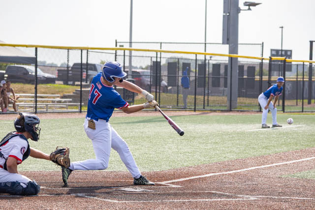 A baseball player in a blue jersey with the number 17 swings a bat to hit a pitch while a catcher in a white jersey crouches behind home plate. The scene takes place on a well-maintained turf baseball field on a bright sunny day.