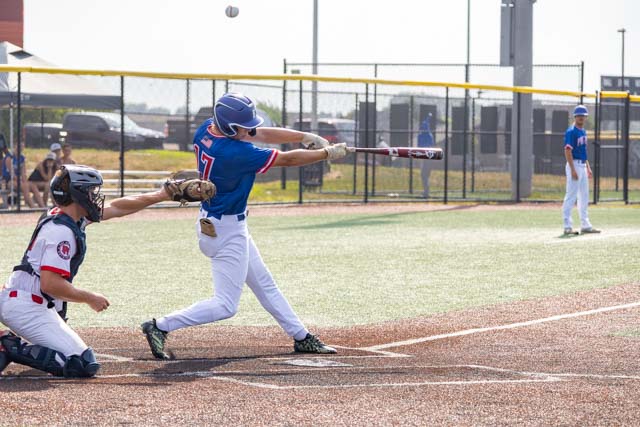 A baseball player in a blue jersey swings at a pitch during a game, while a catcher in a black and white uniform crouches behind home plate.