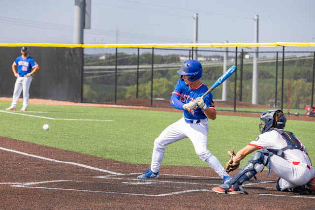 A baseball player prepares to hit a pitch while a catcher waits behind home plate, with another player in the background.