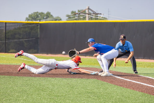 A baseball player in a red uniform is diving towards first base while another player in blue attempts to catch the ball with a glove. An umpire watches the play from the side.