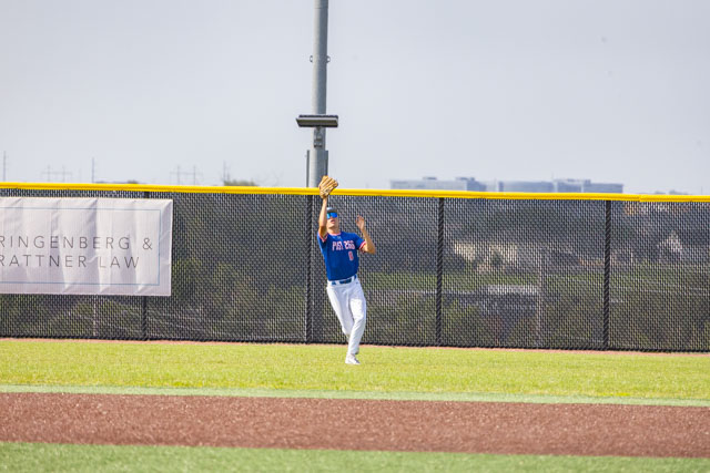 A baseball player in a blue jersey and white pants catching a fly ball in the outfield under a clear sky.
