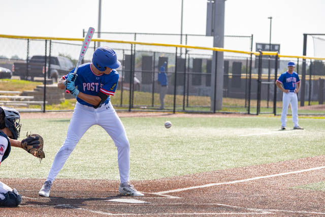 A young baseball player in a blue uniform is about to swing at a pitched ball while another player stands on the field in the background.