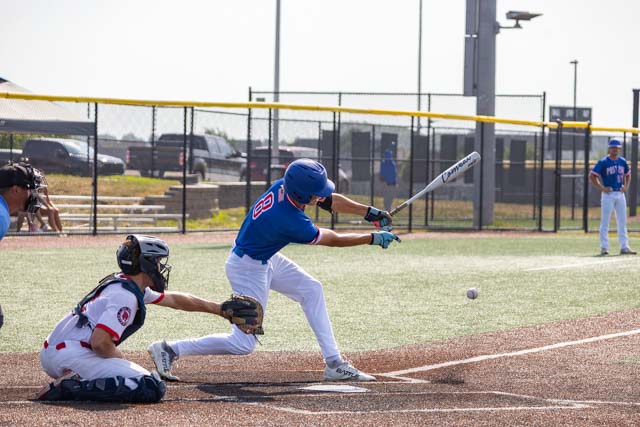 A baseball player in a blue jersey swings at a pitch while a catcher in a black and red uniform crouches behind home plate.