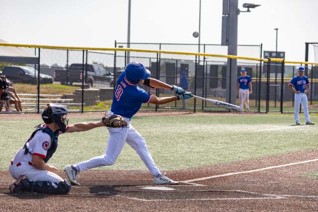 A baseball player swings a bat to hit a pitch while a catcher prepares to catch the ball. The scene takes place on a baseball field with teammates in the background.