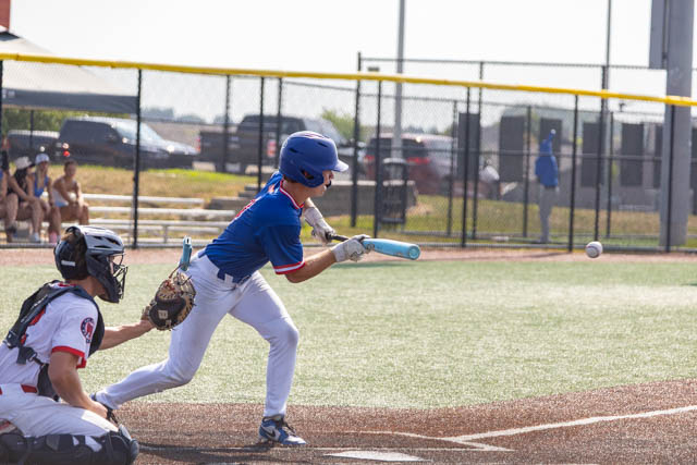 A young baseball player in a blue jersey swings a bat at a pitched ball while a catcher in a black and red uniform squats behind home plate.