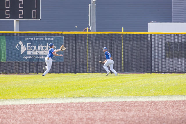 Two baseball players in blue jerseys are in the outfield, one is catching a fly ball while the other runs towards him. A scoreboard and a fenced area are visible in the background.
