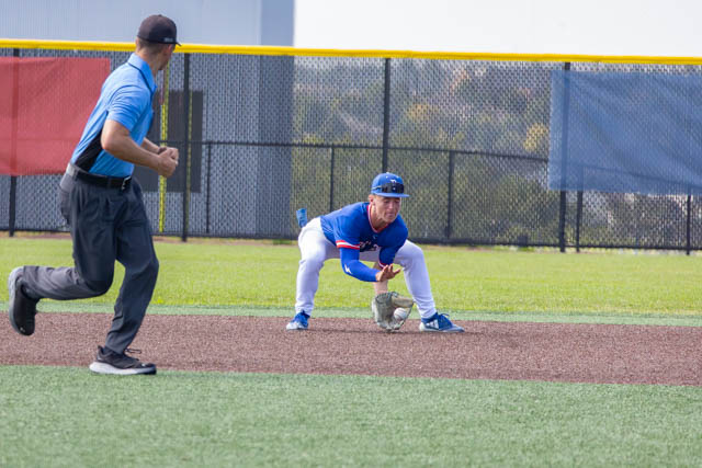 A baseball player in a blue uniform prepares to field a ground ball on a diamond, while an umpire in a blue and black outfit stands nearby.