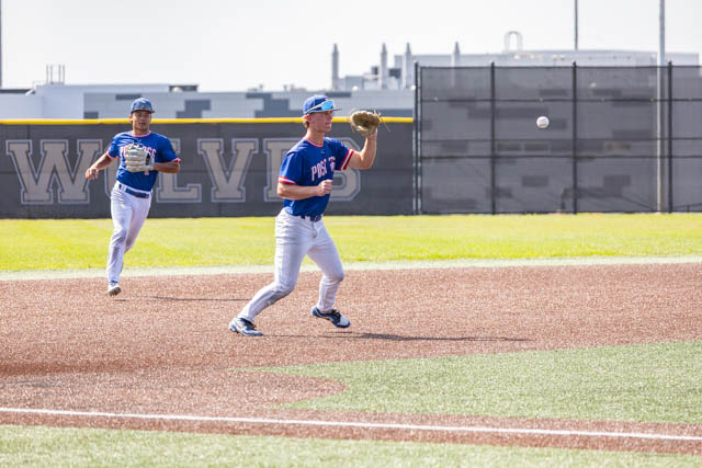 A baseball player in a blue jersey and white pants is catching a ball on the field, while another player in the background is running.