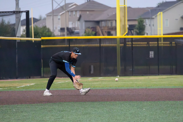 A baseball player crouching down to field a ground ball on a baseball field, with houses in the background.