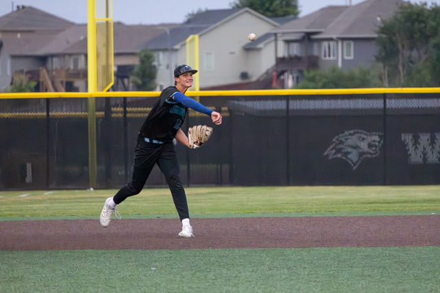 A baseball player in a black uniform fields a ball on a sunny day, preparing to throw from the infield.