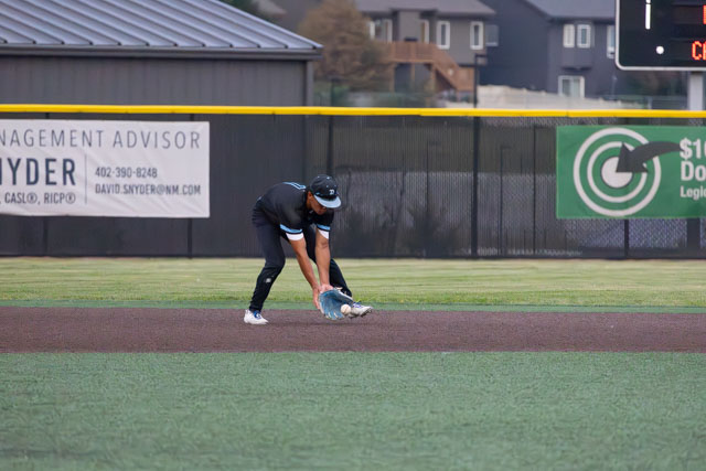 A baseball player crouches down on the field, reaching to catch a ground ball during a game.