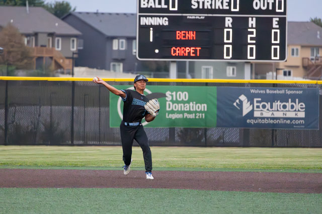 A young baseball player in black uniform fields the ball on a baseball diamond, with a scoreboard displaying the score in the background.