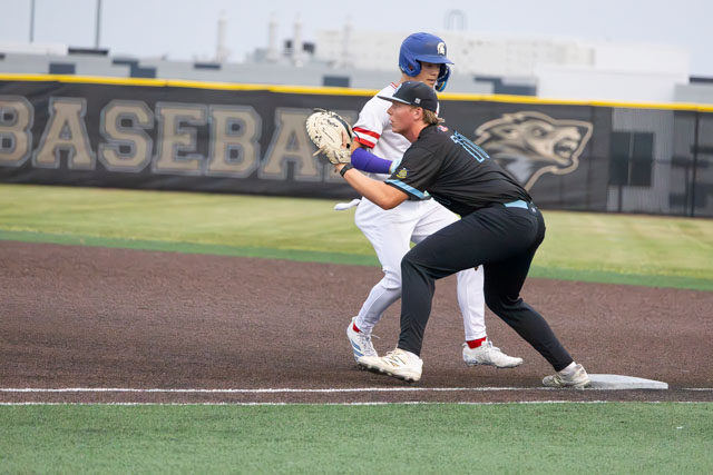 A baseball player in a blue helmet and white uniform is running towards first base while another player in a black uniform is positioned in a defensive stance at first base.