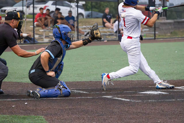 A baseball player swings at a pitch while a catcher prepares to catch the ball, with spectators in the background.