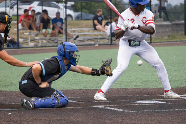 A baseball catcher in a blue helmet and chest protector reaches for a pitch while a batter, dressed in a white uniform with red accents, prepares to swing at the ball.