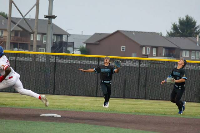 Two young baseball players attempting to catch a fly ball during a game, with a player running towards first base in the foreground.