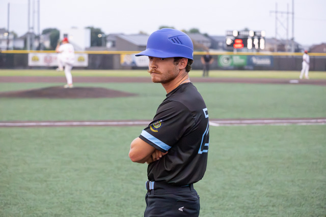 A baseball player in a black uniform with a blue cap stands with arms crossed, observing a game in the background.