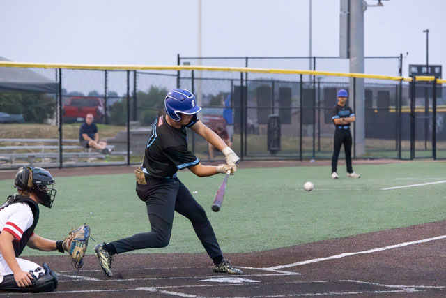 A baseball player swings a bat to hit a ball on a field, with a catcher in position behind home plate and other players in the background.